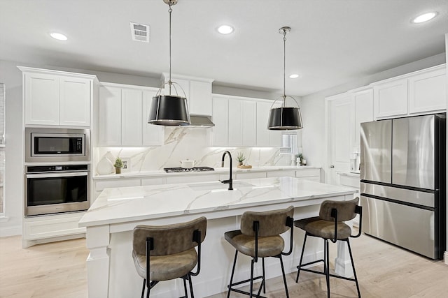 kitchen with hanging light fixtures, a kitchen island with sink, white cabinetry, light wood-type flooring, and appliances with stainless steel finishes