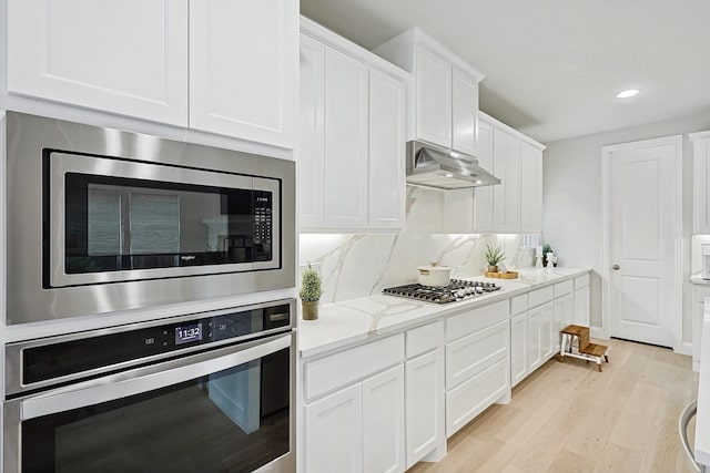 kitchen with light wood-type flooring, appliances with stainless steel finishes, light stone countertops, decorative backsplash, and white cabinets