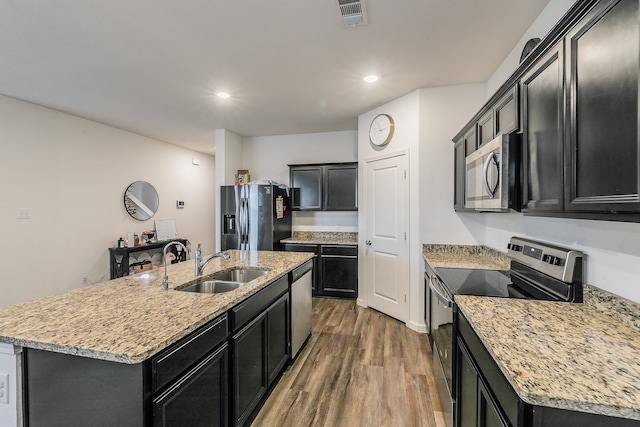 kitchen featuring an island with sink, stainless steel appliances, wood-type flooring, sink, and light stone countertops