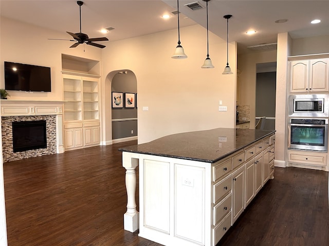 kitchen with a stone fireplace, hanging light fixtures, stainless steel appliances, dark stone countertops, and dark hardwood / wood-style flooring