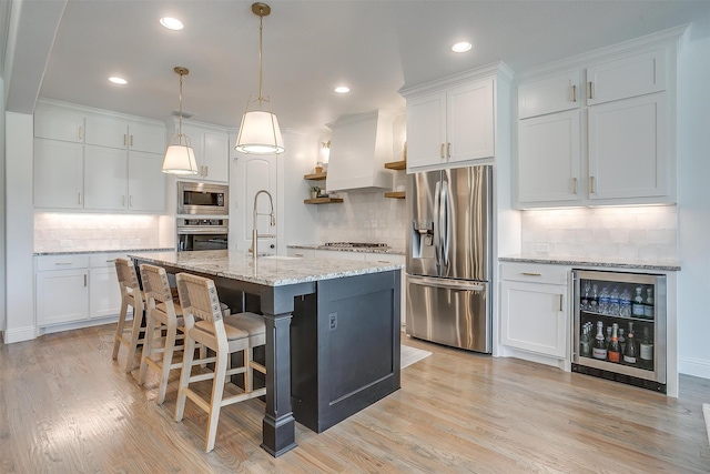 kitchen featuring a kitchen island with sink, hanging light fixtures, stainless steel appliances, wine cooler, and white cabinetry