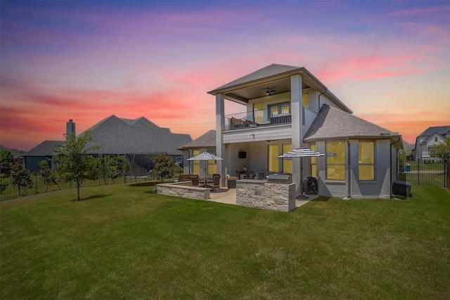 back house at dusk with a yard, a patio, a balcony, exterior kitchen, and ceiling fan