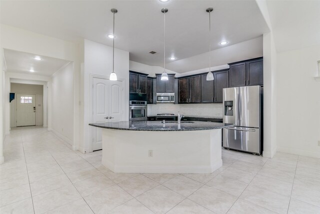 kitchen featuring appliances with stainless steel finishes, dark stone countertops, a center island with sink, hanging light fixtures, and baseboards