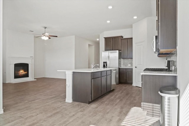 kitchen with light hardwood / wood-style flooring, appliances with stainless steel finishes, an island with sink, ceiling fan, and decorative backsplash