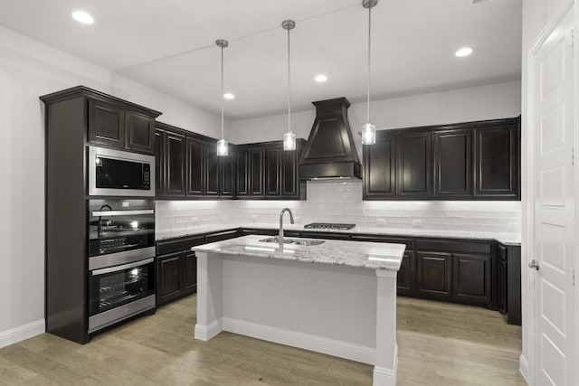 kitchen featuring light hardwood / wood-style floors, sink, premium range hood, a kitchen island with sink, and pendant lighting