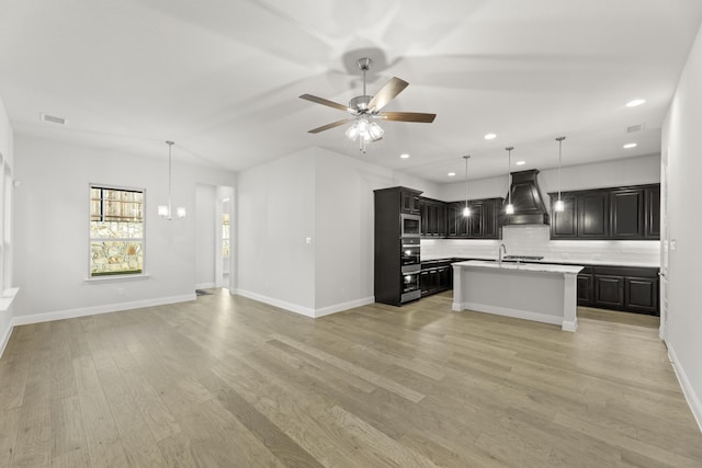 kitchen with a center island with sink, appliances with stainless steel finishes, tasteful backsplash, custom exhaust hood, and light hardwood / wood-style flooring