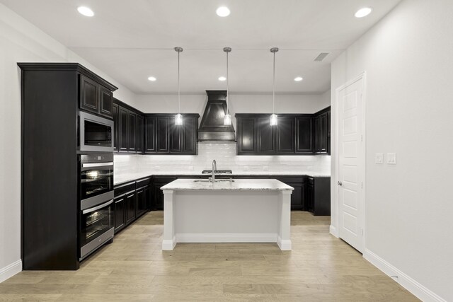 kitchen with an island with sink, custom range hood, backsplash, light wood-type flooring, and decorative light fixtures