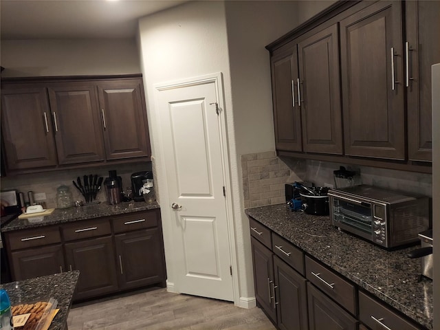 kitchen featuring dark brown cabinets, light wood-type flooring, tasteful backsplash, and dark stone counters