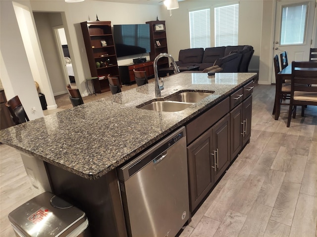 kitchen with dark stone counters, sink, stainless steel dishwasher, light wood-type flooring, and an island with sink