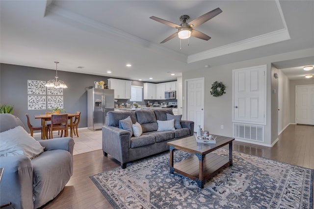 living room with ornamental molding, light hardwood / wood-style flooring, a tray ceiling, and ceiling fan with notable chandelier