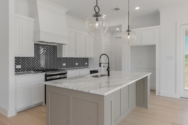 kitchen featuring hanging light fixtures, a center island with sink, light wood-type flooring, premium range hood, and light stone counters