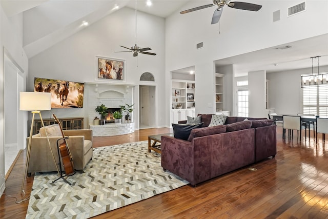 living room with high vaulted ceiling, wood-type flooring, a brick fireplace, ceiling fan with notable chandelier, and built in features