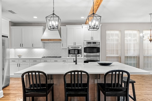 kitchen featuring white cabinetry, a center island with sink, appliances with stainless steel finishes, light hardwood / wood-style flooring, and premium range hood