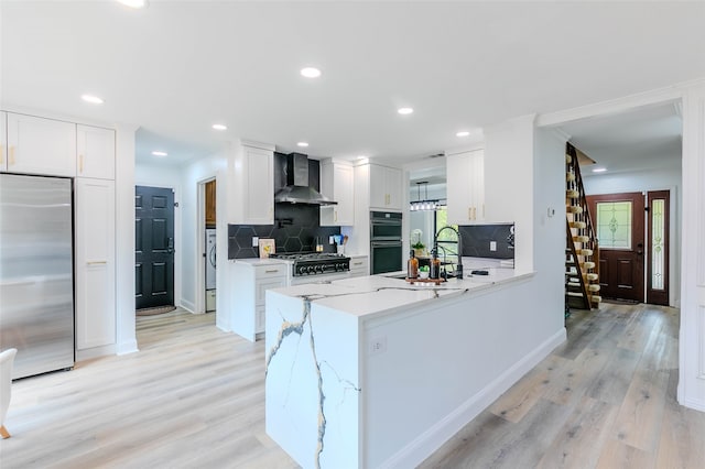 kitchen featuring wall chimney range hood, light hardwood / wood-style flooring, kitchen peninsula, white cabinets, and appliances with stainless steel finishes