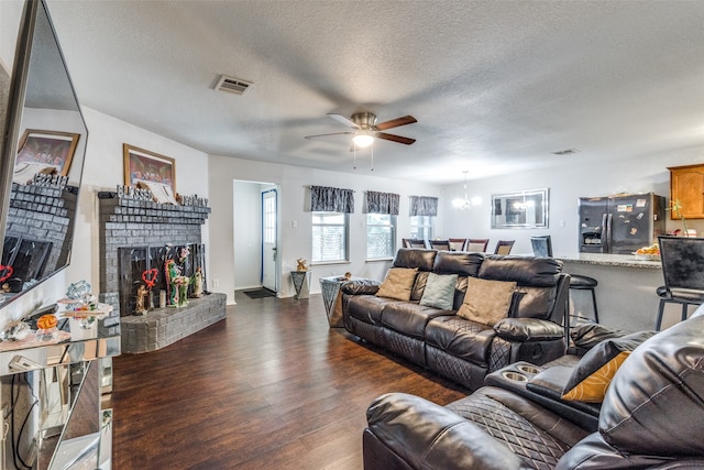 living room featuring ceiling fan with notable chandelier, a textured ceiling, a brick fireplace, and dark hardwood / wood-style floors