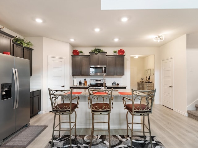 kitchen featuring dark brown cabinetry, light stone countertops, stainless steel appliances, backsplash, and an island with sink