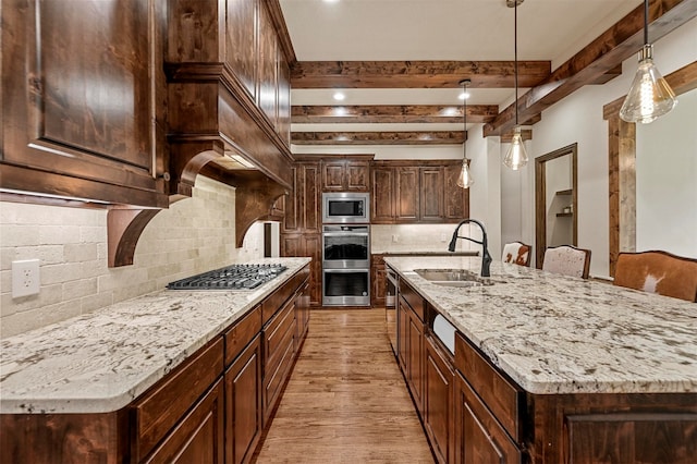 kitchen featuring a large island with sink, sink, light hardwood / wood-style floors, stainless steel appliances, and beamed ceiling