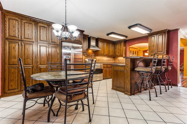 kitchen with wall chimney range hood, crown molding, light tile patterned floors, stainless steel appliances, and a chandelier