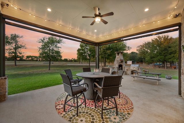 patio terrace at dusk with ceiling fan, an outdoor stone fireplace, and a lawn