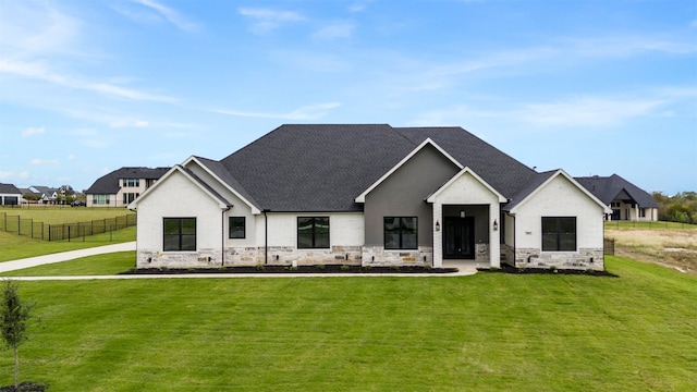 modern farmhouse with a shingled roof, a front yard, stone siding, and fence