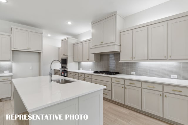 kitchen featuring appliances with stainless steel finishes, light hardwood / wood-style floors, custom range hood, decorative backsplash, and a center island with sink