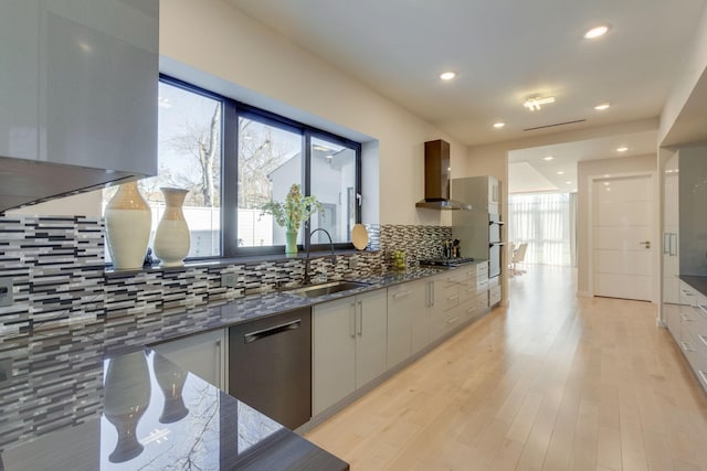 kitchen with dark stone counters, wall chimney range hood, sink, decorative backsplash, and light wood-type flooring