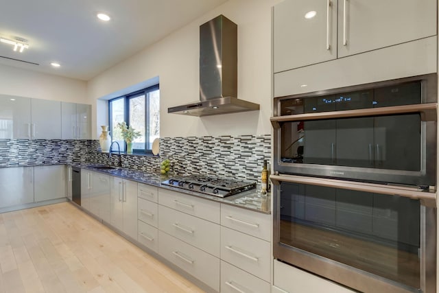 kitchen featuring appliances with stainless steel finishes, tasteful backsplash, wall chimney exhaust hood, sink, and dark stone countertops