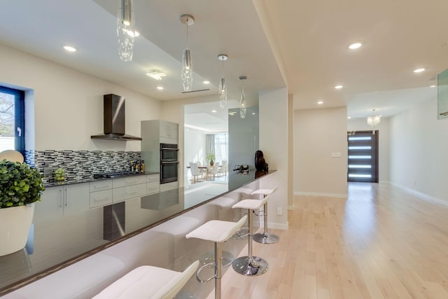 kitchen with light wood-type flooring, backsplash, wall chimney exhaust hood, black double oven, and white cabinetry