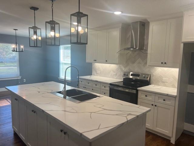 kitchen with wall chimney exhaust hood, decorative light fixtures, stainless steel electric range, white cabinets, and an island with sink