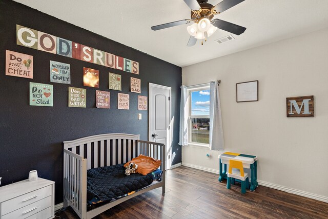 bedroom with dark wood-type flooring, a crib, and ceiling fan