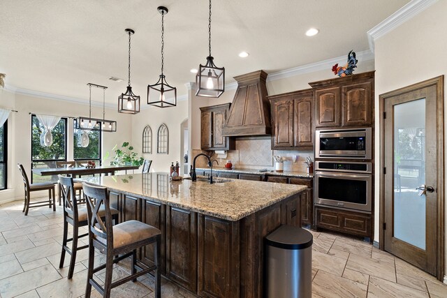 kitchen with appliances with stainless steel finishes, light stone countertops, custom exhaust hood, decorative light fixtures, and an island with sink