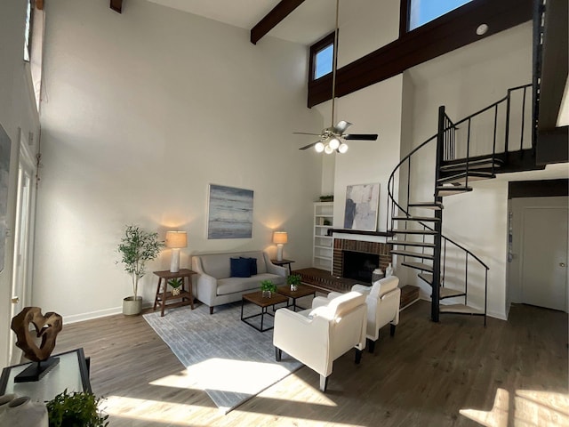 living room featuring a towering ceiling, a brick fireplace, ceiling fan, wood-type flooring, and beam ceiling