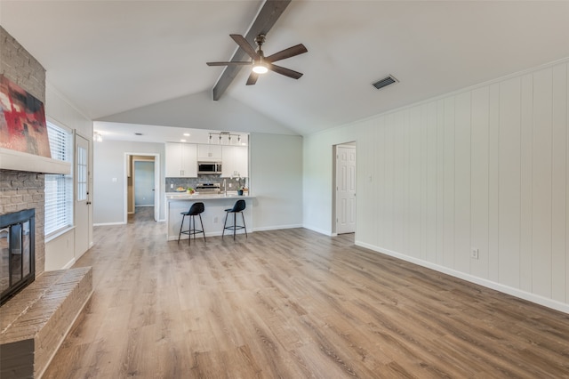 unfurnished living room with light hardwood / wood-style flooring, a brick fireplace, lofted ceiling with beams, and ceiling fan