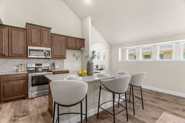 kitchen with appliances with stainless steel finishes, high vaulted ceiling, light wood-type flooring, and an island with sink