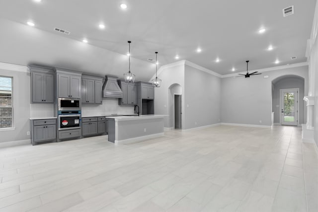 kitchen with gray cabinetry, hanging light fixtures, stainless steel appliances, custom exhaust hood, and a kitchen island with sink