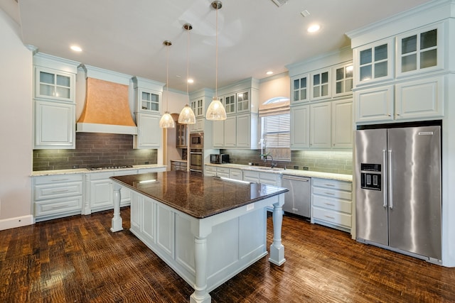 kitchen featuring stainless steel appliances, a kitchen island, custom range hood, pendant lighting, and dark stone countertops