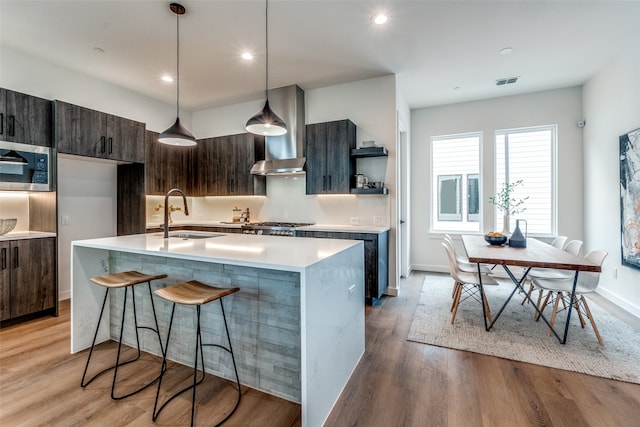 kitchen with hanging light fixtures, a kitchen island with sink, light wood-type flooring, dark brown cabinetry, and sink