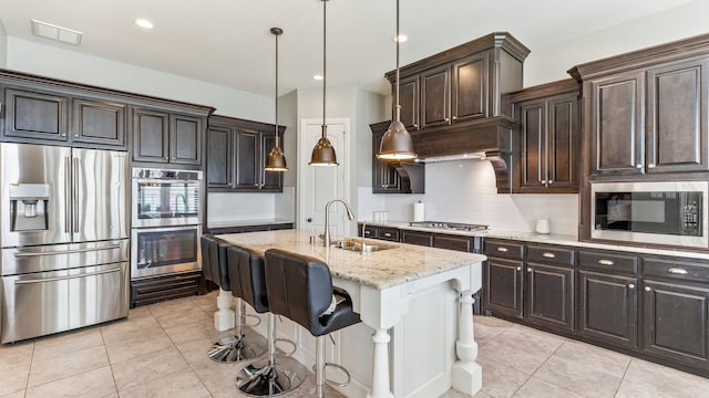 kitchen with sink, a kitchen island with sink, light stone countertops, appliances with stainless steel finishes, and decorative light fixtures