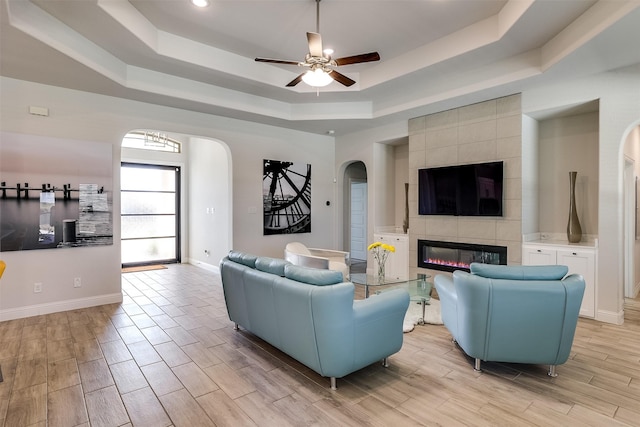 living room featuring ceiling fan with notable chandelier, a raised ceiling, and a tiled fireplace