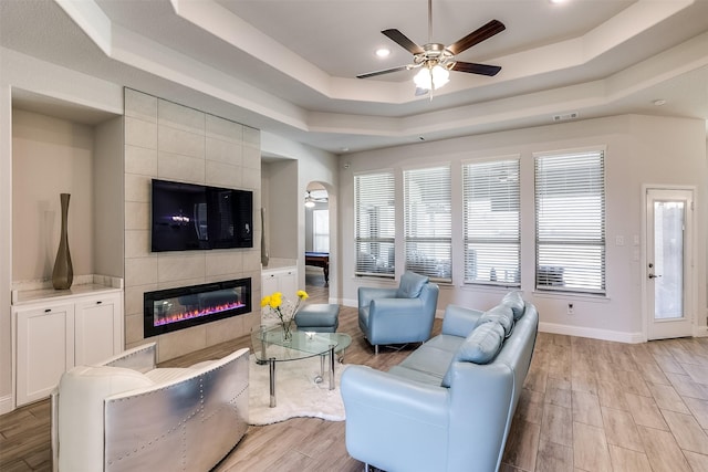 living area featuring a tile fireplace, a ceiling fan, baseboards, light wood-style floors, and a tray ceiling