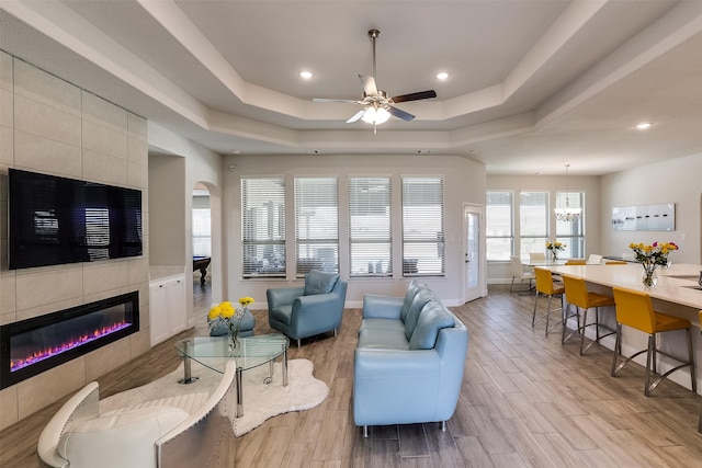 living room with ceiling fan with notable chandelier, a tray ceiling, and a tiled fireplace