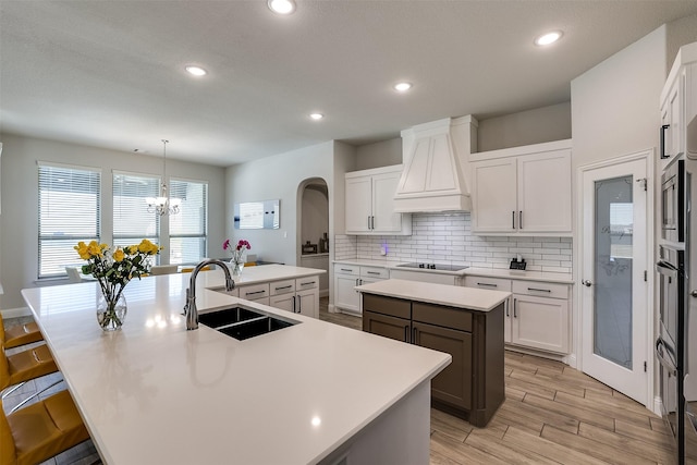 kitchen with an island with sink, black electric stovetop, custom range hood, pendant lighting, and sink