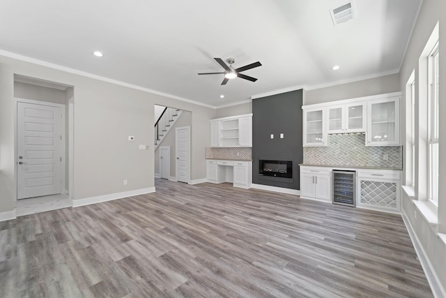 unfurnished living room featuring a large fireplace, beverage cooler, ornamental molding, ceiling fan, and light wood-type flooring