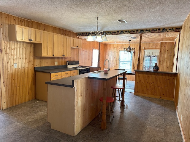 kitchen with white gas range, a center island with sink, an inviting chandelier, wood walls, and decorative light fixtures