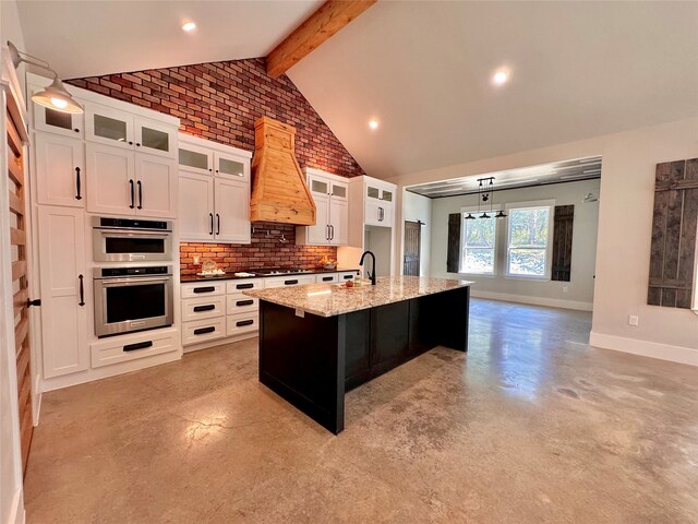 kitchen featuring vaulted ceiling with beams, custom range hood, stainless steel appliances, a center island with sink, and white cabinets