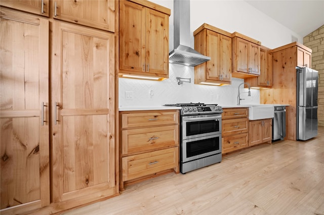 kitchen with wall chimney range hood, appliances with stainless steel finishes, sink, lofted ceiling, and light hardwood / wood-style flooring