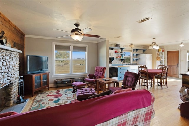 living room with light wood-type flooring, a stone fireplace, ceiling fan, and crown molding