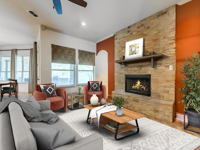 living room with light hardwood / wood-style floors, ornamental molding, a stone fireplace, and ceiling fan