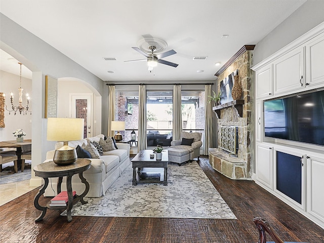 living room featuring dark wood-type flooring, a stone fireplace, and ceiling fan with notable chandelier