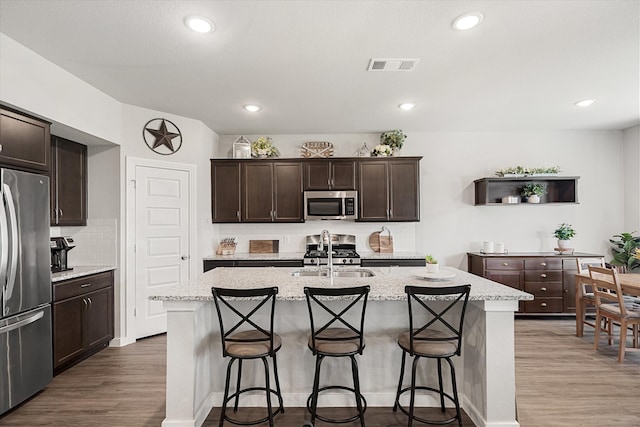 kitchen with an island with sink, appliances with stainless steel finishes, light stone countertops, dark wood-type flooring, and sink